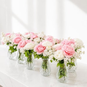 May include: A series of six clear, ribbed glass vases, each containing a floral arrangement of soft pink and white ranunculus flowers accented with small white filler blossoms and green leaves, arranged in a line on a white surface.