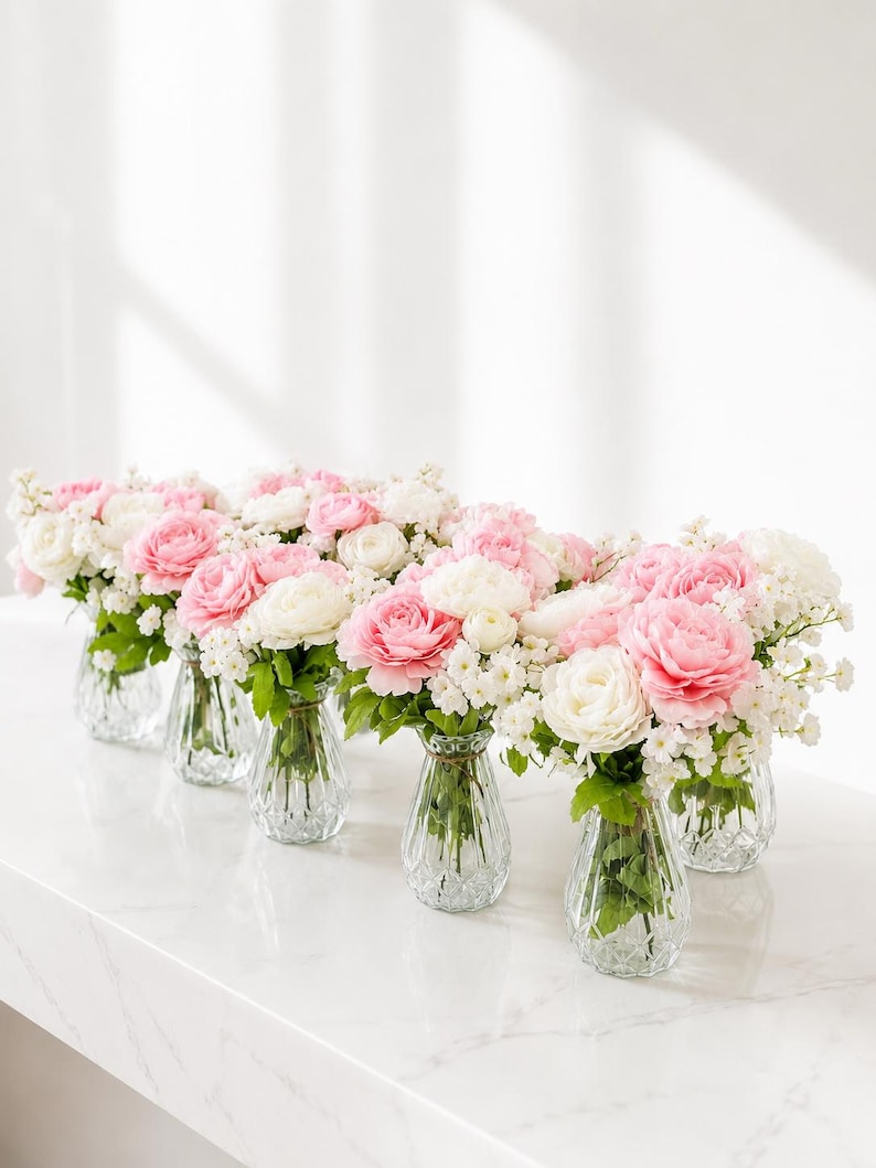 May include: A series of six clear, ribbed glass vases, each containing a floral arrangement of soft pink and white ranunculus flowers accented with small white filler blossoms and green leaves, arranged in a line on a white surface.