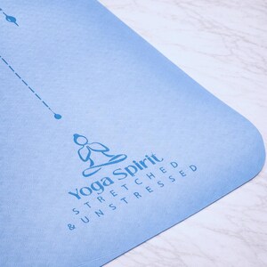 Pu&ograve; includere: Un tappetino da yoga azzurro con angoli arrotondati. Il tappetino presenta il testo "Yoga Spirit" e le parole "Stretched & Unstressed" in blu. Sono visibili anche una linea tratteggiata e un piccolo cerchio.