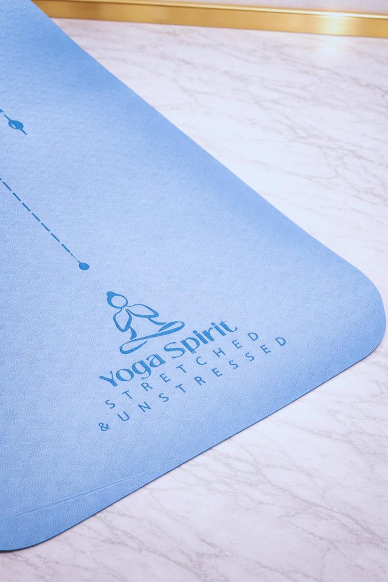 Pu&ograve; includere: Un tappetino da yoga azzurro con angoli arrotondati. Il tappetino presenta il testo "Yoga Spirit" e le parole "Stretched & Unstressed" in blu. Sono visibili anche una linea tratteggiata e un piccolo cerchio.