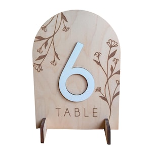 May include: Wooden table number six with a light blue number and floral design. The word "TABLE" is printed at the bottom. The table number is supported by two wooden stands.