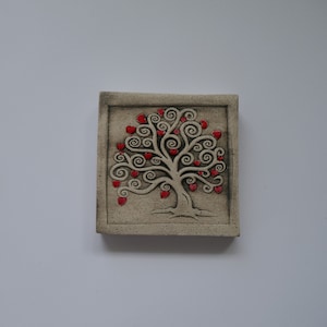 Könnte beinhalten: Eine quadratische, beige Keramikfliese mit einem Baumdesign und roten herzförmigen Blättern. Der Baum hat ein detailliertes, wirbelndes Muster. Die Fliese ist vor einem weißen Hintergrund platziert und zeigt ein dekoratives, handgefertigtes Objekt.