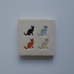 Könnte beinhalten: Ein quadratischer, cremefarbener Fliesenstein zeigt vier Katzensilhouetten in Schwarz, Blau, Orange und Beige. Die Katzen sind gleichmäßig verteilt und in die Oberfläche der Fliese geschnitzt. Die Fliese hat ein strukturiertes Aussehen.