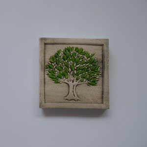 Pode incluir: Azulejo de cerâmica bege quadrado com um relevo detalhado de uma árvore. A árvore tem um tronco cinza claro e folhas verdes vibrantes. O azulejo está emoldurado por uma borda em relevo.