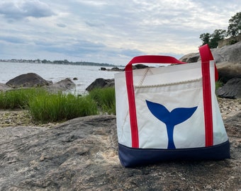 May include: White tote bag with a blue whale tail design. The bag has red handles and a navy blue bottom. The bag is made of a sailcloth material and is sitting on a rock near the ocean.