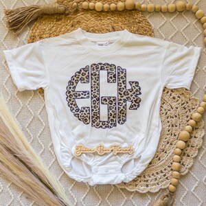 May include: White baby bodysuit with a leopard print monogram design. The monogram features the letters "EGH" in a circular pattern with a scalloped edge. The bodysuit has short sleeves and a snap closure. The wooden tag reads "Juice Box Threads".
