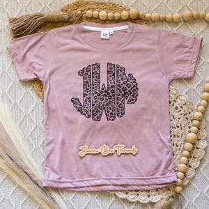 May include: A mauve-colored t-shirt featuring a leopard print monogram of the letters JWM. The shirt has a crew neck and short sleeves. The words "Juice Box Threads" are printed on a wooden sign below the monogram.
