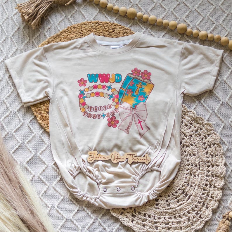 May include: A beige baby onesie with a colorful graphic featuring a book, rosary, and the words "WWJD", "Faith", and "Pray to God". The design includes pink flowers and a pink bow. The text "Juice Box Threads" is printed at the bottom.
