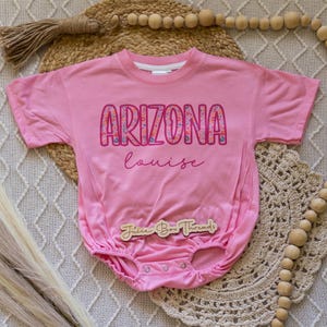 Pu&ograve; includere: Un body rosa per neonati con la scritta "ARIZONA" in un motivo floreale e il nome "louise" in corsivo. Il body ha maniche corte e bottoni a pressione sul fondo. &Egrave; visibile anche un'etichetta di legno con "Juice Box Threads".