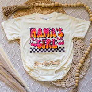 May include: Cream-colored baby bodysuit with the text "MAMA'S GIRL" in a retro font with pink and orange flames, cherries, and a checkered pattern. The bodysuit has short sleeves and snap closures.