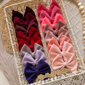 May include: A collection of 10 pairs of handmade hair bows in various shades of red, pink, purple, and gray. The bows are arranged in a white wicker basket. The bows are made of a soft, textured fabric and are perfect for adding a touch of whimsy to any outfit.
