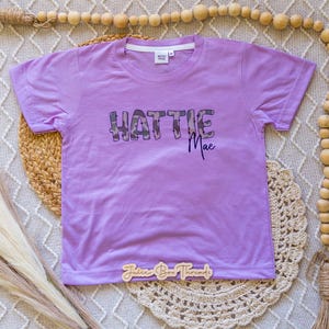 Puede incluir: Una camiseta de manga corta de color morado claro con el texto HATTIE MAE impreso en la parte delantera. La palabra HATTIE est&aacute; en una fuente grande y cuadrada con patr&oacute;n de camuflaje, y la palabra Mae en cursiva morada oscura.