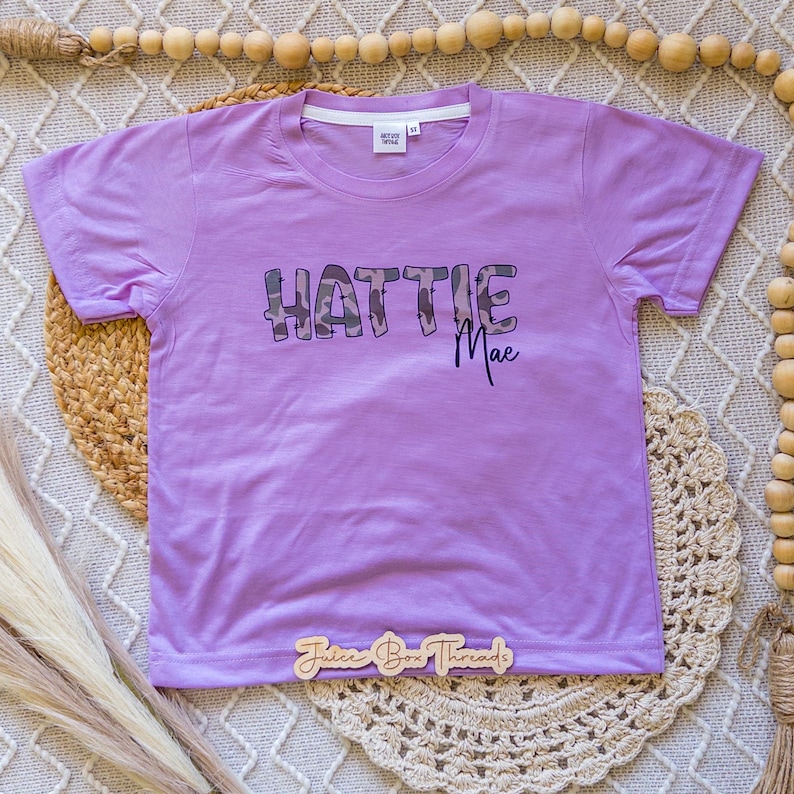 Puede incluir: Una camiseta de manga corta de color morado claro con el texto HATTIE MAE impreso en la parte delantera. La palabra HATTIE est&aacute; en una fuente grande y cuadrada con patr&oacute;n de camuflaje, y la palabra Mae en cursiva morada oscura.