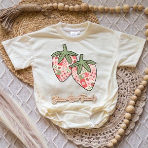 May include: Cream-colored baby onesie featuring a strawberry design. The strawberries have a patchwork pattern in pink, green, and white. The text "Juice Box Threads" is printed on a wooden tag below the onesie.