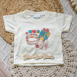 May include: Cream-colored short-sleeved t-shirt with a colorful graphic. The graphic includes a beaded bracelet, a book with a cross, and the text "WWJD", "Faith", and "Pray+God". The shirt also has the text "Juice Box Threads".
