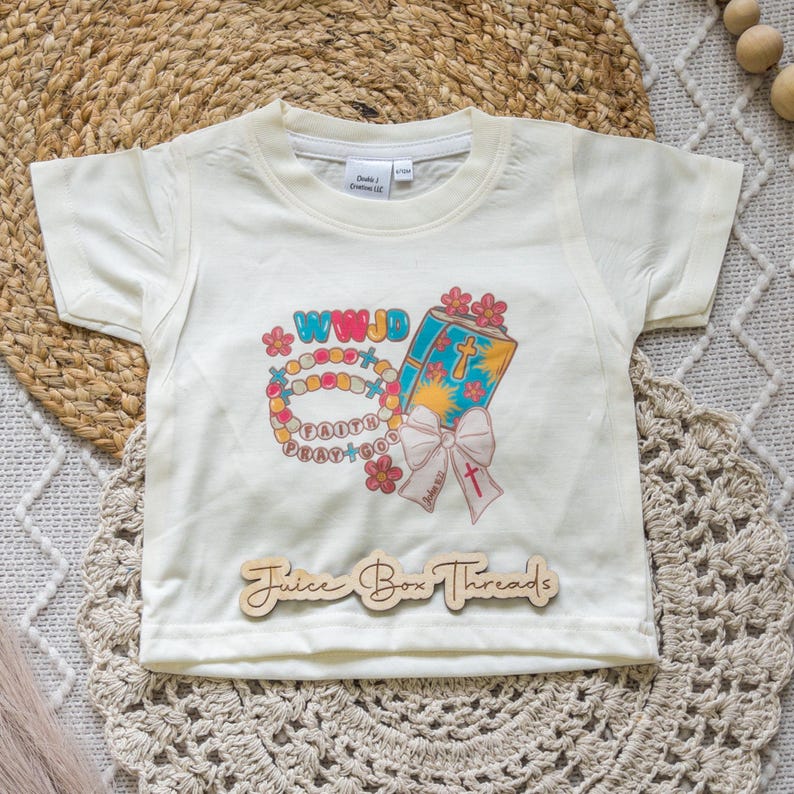 May include: Cream-colored short-sleeved t-shirt with a colorful graphic. The graphic includes a beaded bracelet, a book with a cross, and the text "WWJD", "Faith", and "Pray+God". The shirt also has the text "Juice Box Threads".
