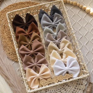 May include: A wicker basket filled with a variety of handmade hair bows in neutral colors, including brown, beige, and white. The bows are arranged in rows and are all made from a soft, textured fabric. The basket is sitting on a woven rug. The text "Double J Creations" is visible on a wooden tag.