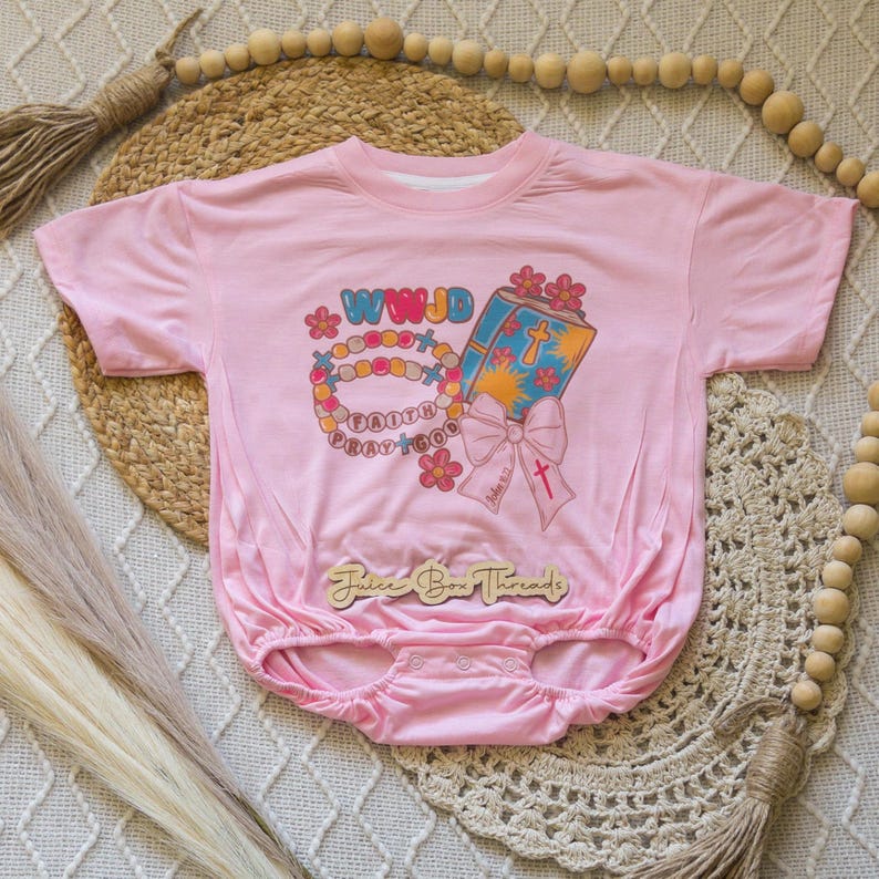 May include: Pink baby onesie with a graphic print featuring a rosary, a book with a cross, and the text "WWJD Faith Pray God." The onesie has short sleeves and a round neckline. The brand name "Juice Box Threads" is also visible.