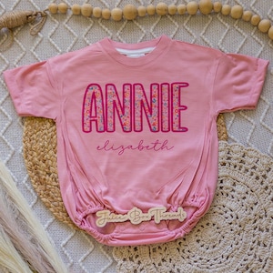 Pu&ograve; includere: Un body rosa per neonati con il nome "ANNIE" in grandi lettere colorate. Il nome "Elizabeth" &egrave; scritto in corsivo sotto. Il body ha maniche corte e vita arricciata. Il marchio "Juice Box Threads" &egrave; stampato su un'etichetta di legno.