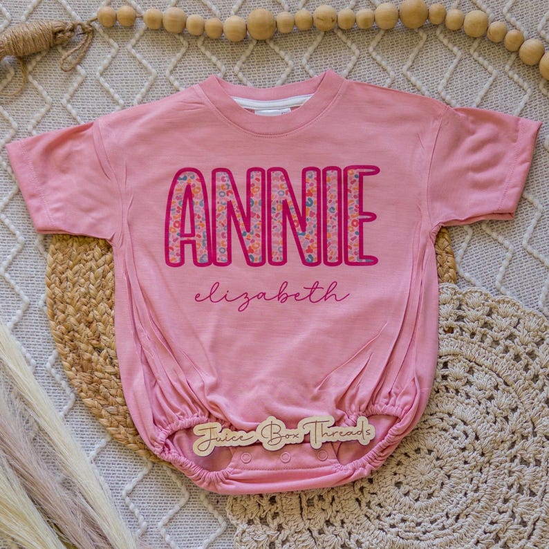 Pu&ograve; includere: Un body rosa per neonati con il nome "ANNIE" in grandi lettere colorate. Il nome "Elizabeth" &egrave; scritto in corsivo sotto. Il body ha maniche corte e vita arricciata. Il marchio "Juice Box Threads" &egrave; stampato su un'etichetta di legno.
