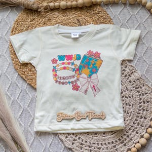 May include: Cream-colored toddler t-shirt with a colorful graphic. The graphic includes the text "WWJD", a rosary, a book, and a pink bow. The words "Faith Pray God" are also visible. The shirt has a Juice Box Threads wooden tag.