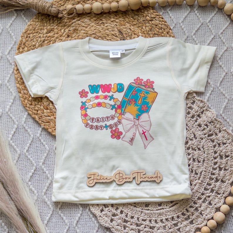 May include: Cream-colored toddler t-shirt with a colorful graphic. The graphic includes the text "WWJD", a rosary, a book, and a pink bow. The words "Faith Pray God" are also visible. The shirt has a Juice Box Threads wooden tag.