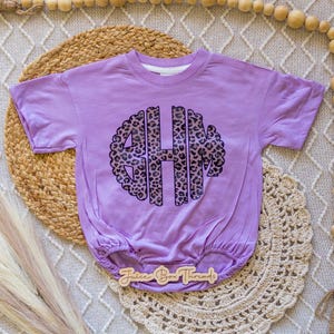 May include: A lavender baby romper with a large monogram design in a leopard print pattern. The romper has short sleeves and a gathered bottom. The text "Juice Box Threads" is visible on a wooden tag.