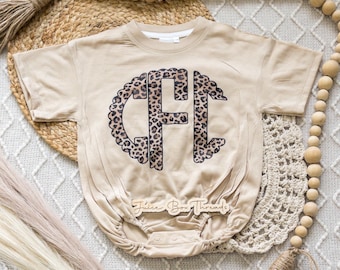 May include: Tan baby romper with a large leopard print monogram. The monogram is in the center of the romper and is surrounded by a scalloped edge. The text "Juice Box Threads" is on a wooden tag at the bottom of the romper.