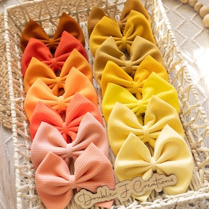 May include: A basket filled with a variety of handmade hair bows in shades of orange, yellow, and peach. The bows are arranged in a gradient pattern, with the darkest orange at the bottom and the lightest peach at the top. The bows are made of a soft, textured fabric and have a double-loop design. The basket is made of woven wicker and has a natural brown color. The text "Double J Creations" is visible on a wooden tag in the lower right corner of the image.