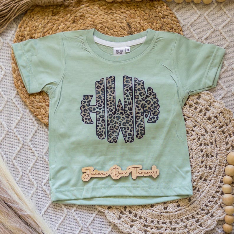 May include: A light green short-sleeved t-shirt with a leopard print monogram design. The shirt has the text "Juice Box Threads" on a wooden tag. The shirt is size 2T.