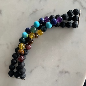 K&ouml;nnte beinhalten: Ein mehrreihiges Armband mit bunten Edelsteinperlen, darunter T&uuml;rkis, Amethyst und Tigerauge, abwechselnd mit schwarzen Lavasteinperlen. Das Armband wird durch silberfarbene Metallst&auml;be zusammengehalten. Die Perlen haben einen Durchmesser von ca. 1 cm.