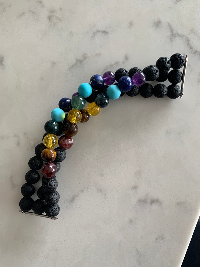 K&ouml;nnte beinhalten: Ein mehrreihiges Armband mit bunten Edelsteinperlen, darunter T&uuml;rkis, Amethyst und Tigerauge, abwechselnd mit schwarzen Lavasteinperlen. Das Armband wird durch silberfarbene Metallst&auml;be zusammengehalten. Die Perlen haben einen Durchmesser von ca. 1 cm.