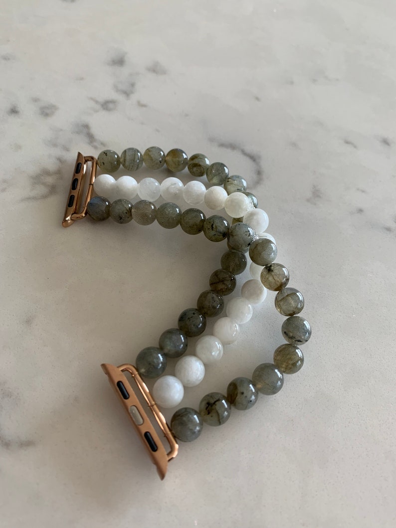 K&ouml;nnte beinhalten: Ein dreireihiges Armband f&uuml;r die Apple Watch mit grauen und wei&szlig;en Perlen. Das Armband hat einen ros&eacute;goldenen Verschluss.