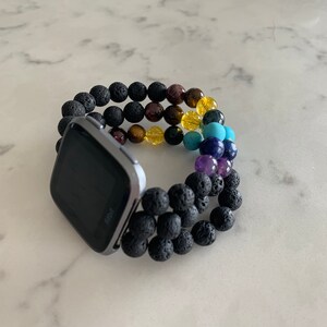 K&ouml;nnte beinhalten: Eine Fitbit-Smartwatch mit schwarzem Bildschirm ist an einem Perlenarmband befestigt. Das Armband hat schwarze Lavaperlen und ein Spektrum an farbigen Edelsteinen, darunter Lila, Blau, Gelb und Braun. Die Uhr und das Armband liegen auf einer wei&szlig;en Marmoroberfl&auml;che.