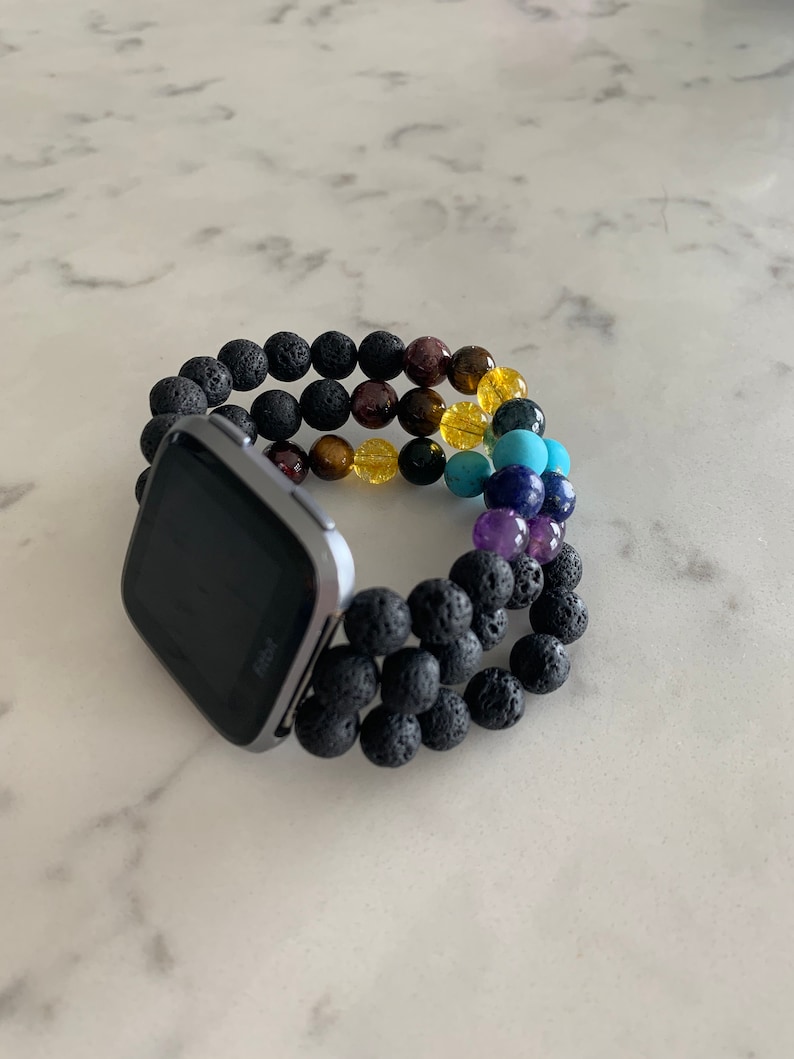 K&ouml;nnte beinhalten: Eine Fitbit-Smartwatch mit schwarzem Bildschirm ist an einem Perlenarmband befestigt. Das Armband hat schwarze Lavaperlen und ein Spektrum an farbigen Edelsteinen, darunter Lila, Blau, Gelb und Braun. Die Uhr und das Armband liegen auf einer wei&szlig;en Marmoroberfl&auml;che.