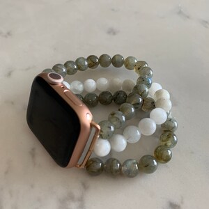 K&ouml;nnte beinhalten: Eine goldene Apple Watch mit schwarzem Zifferblatt und einem Perlenarmband aus grauen und wei&szlig;en Steinen.