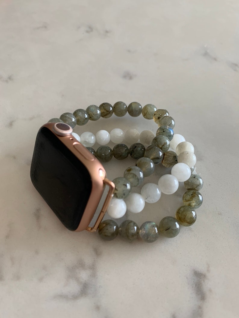 K&ouml;nnte beinhalten: Eine goldene Apple Watch mit schwarzem Zifferblatt und einem Perlenarmband aus grauen und wei&szlig;en Steinen.