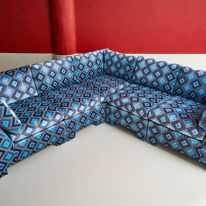 May include: A large, corner sofa with a geometric diamond pattern in shades of blue, gray, and white. The sofa has a modern design with angled arms and backrests. The background is a red wall.