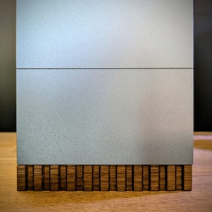 Puede incluir: Un objeto rectangular con una sección superior de color plateado mate y liso, dividida por una línea horizontal, que descansa sobre una base compuesta por numerosas láminas de madera de color marrón oscuro, verticales y texturizadas.