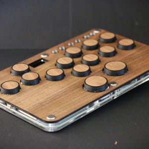 May include: A rectangular wooden device with multiple round buttons. The device has a clear acrylic border and small silver fasteners. The buttons are light brown with black rims. The device is likely a controller or interface.