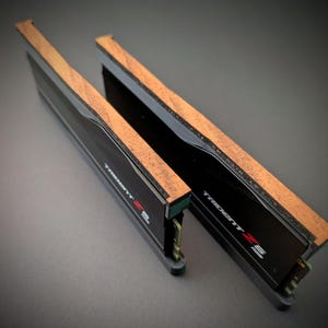 以下が含まれることがあります： 木製のアクセントが付いた2つの黒いコンピューターRAMスティック。「TRIDENT Z」の文字が印刷されています。木製のアクセントは明るい茶色です。背景は濃い灰色です。