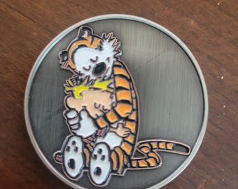 Op de afbeelding: Een ronde metalen speld met een gekleurde emaille afbeelding van een jonge jongen met stekelig geel haar die een grote oranje en zwart gestreepte tijger knuffelt. De achtergrond is van geborsteld grijs metaal met een zilveren rand.