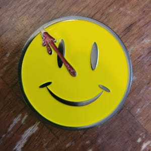 May include: A circular metal pin featuring a bright yellow smiley face with two oval eyes and a curved mouth. A red blood splatter design extends from the top edge of the pin, dripping down across the left eye, creating a stark contrast against the yellow.