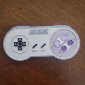 May include: A Super Nintendo video game controller pin featuring a light grey body with a lavender border. It includes a black directional pad, two black buttons labelled Select and Start, and four circular buttons in shades of purple and lavender.