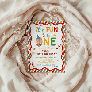 May include: A rectangular birthday invitation card with a white center and a colorful diagonal striped border. The text reads It's FUN to be ONE, Adam's First Birthday, date, time, address, and RSVP details, featuring a photo of a baby in a blue hat.