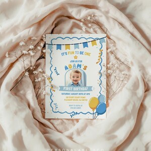 May include: A rectangular first birthday invitation card featuring a scalloped blue border, yellow and blue bunting, and a central photo of a child. The text reads: It's fun to be one, join us for Adam's first birthday, Saturday, August 30th at 4pm.