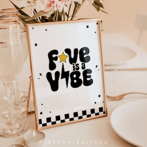 May include: A gold-framed sign with a white background and black text that reads "Fun is a Vibe" with a yellow star and lightning bolt. The sign is on a table with a checkered pattern at the bottom.