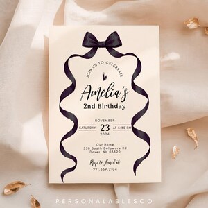 May include: A cream-colored birthday invitation card featuring a black bow at the top with two long, flowing ribbons framing the text. The card reads: Join us to celebrate Amelia's 2nd Birthday, Saturday November 23 2024 at 5:30 PM, 558 South Delaware Rd, Dover, NH 03820.