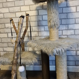 May include: A grey cat tree with two platforms covered in fluffy grey fabric. The tree has a sisal-wrapped post and a dangling toy with a white pom-pom. The tree is in front of a brick wall.
