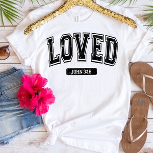 Op de afbeelding: Wit T-shirt met zwarte tekst die "LOVED" in een varsity-stijl lettertype leest. Onder de tekst staat "JOHN 3:16".